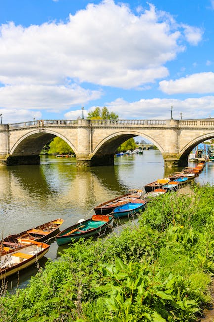 A scenic view of a stone arched bridge crossing a river with a row of small, colorful boats moored along the grassy riverbank in the foreground. The boats have wooden and plastic hulls painted in shades of blue, red, green, and brown, some with canopies or covers. The bridge features classical stone balustrades and lamp posts, with a few trees visible beyond it under a bright blue sky dotted with fluffy white clouds. The river water is calm, reflecting the bridge and sky, and the surrounding environment suggests an outdoor setting suitable for private boat storage or local leisure activities. This scene exemplifies a tranquil waterside area that could be part of a private or independent rubbish removal or waste management scenario near a natural or urban environment, consistent with alternative waste handling where boats or outdoor storage might be temporarily used. The lush green vegetation along the bank adds to the natural atmosphere, and the overall composition emphasizes open-air space typical of a location where outdoor rubbish collection or clearance activities might be undertaken through separate, independent services such as [COMPANY_NAME].