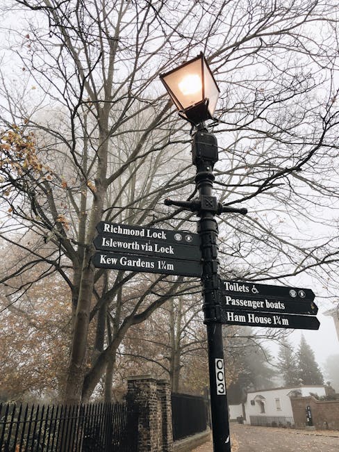 A tall black street signpost with multiple directional signs attached, located beside a pathway in a park area. The signpost features a classic lantern-style streetlight at the top, illuminating the scene with a warm glow. Surrounding the signpost are leafless trees with intricate, spindly branches extending into a foggy, overcast sky, creating a moody atmosphere. The directional signs display white text on black backgrounds, indicating distances and directions to locations such as Richmond Lock, Isleworth via Lock, Kew Gardens 1/4 mile, toilets and passenger boats, and Ham House 1/4 mile. The base of the signpost is anchored in the ground amidst a black metal fence, with a brick wall and white building partially visible in the background on the right. The overall scene suggests a quiet, historic park or riverside area, possibly indirectly related to waste or rubbish management through public signage, set in a typical urban environment with a subtle nod to private or local waste management practices.